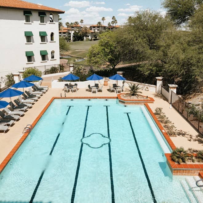 A large outdoor pool with lounge chairs and umbrellas, surrounded by a building and trees.