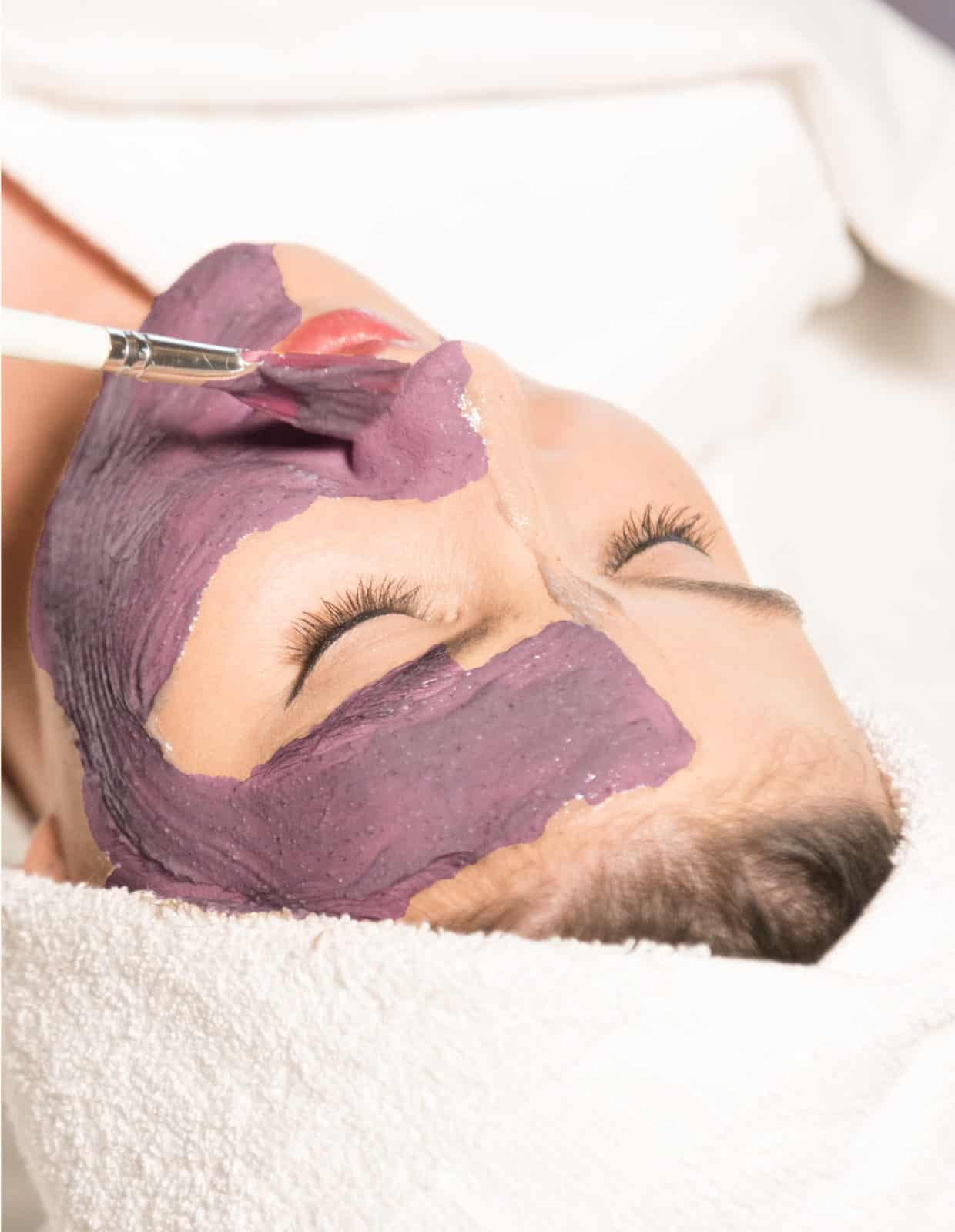 A woman receiving a facial treatment with a purple mask being applied to her face.