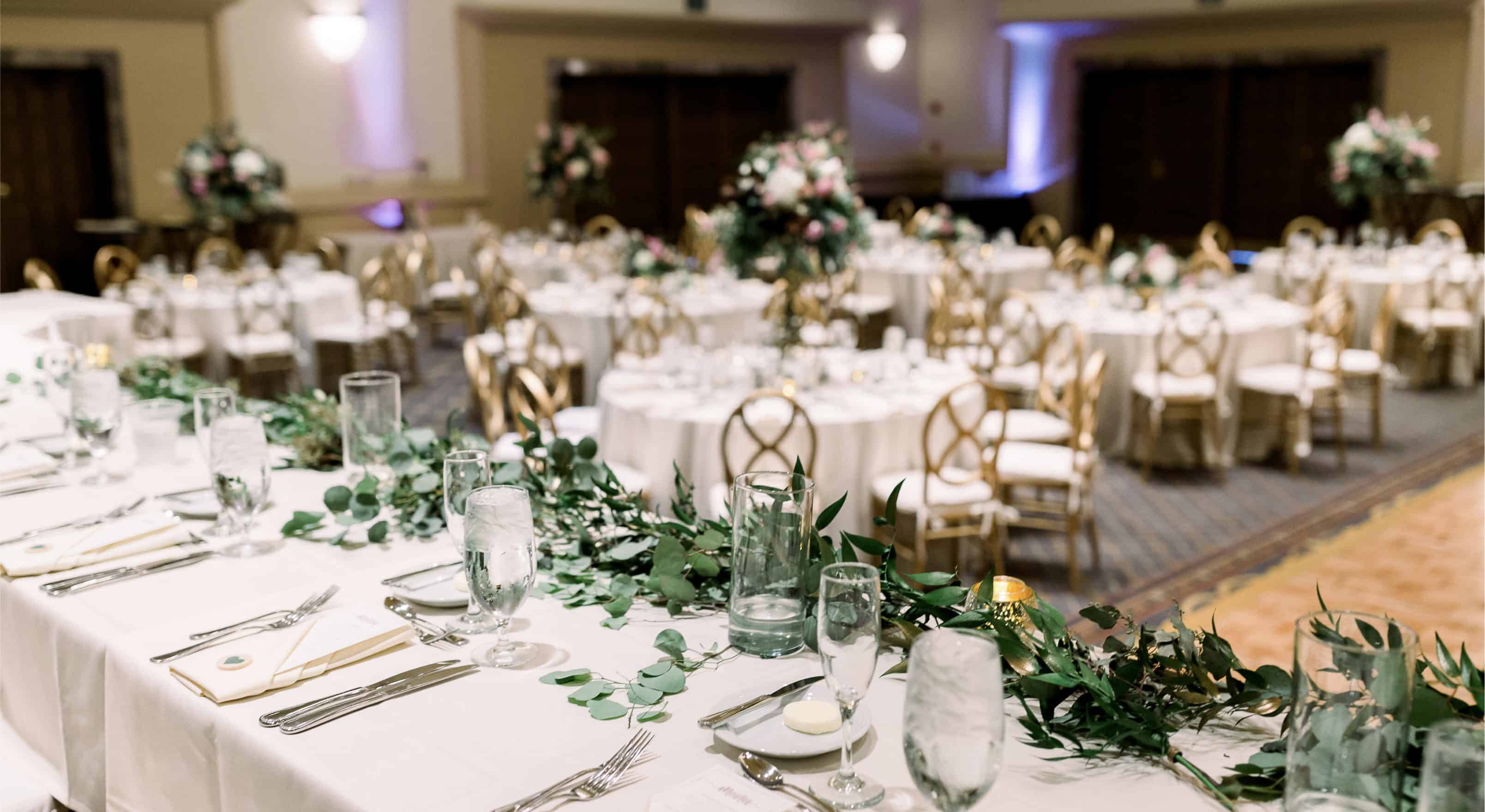 A large, elegantly set dining room with white tablecloths, gold chairs, and greenery garlands.