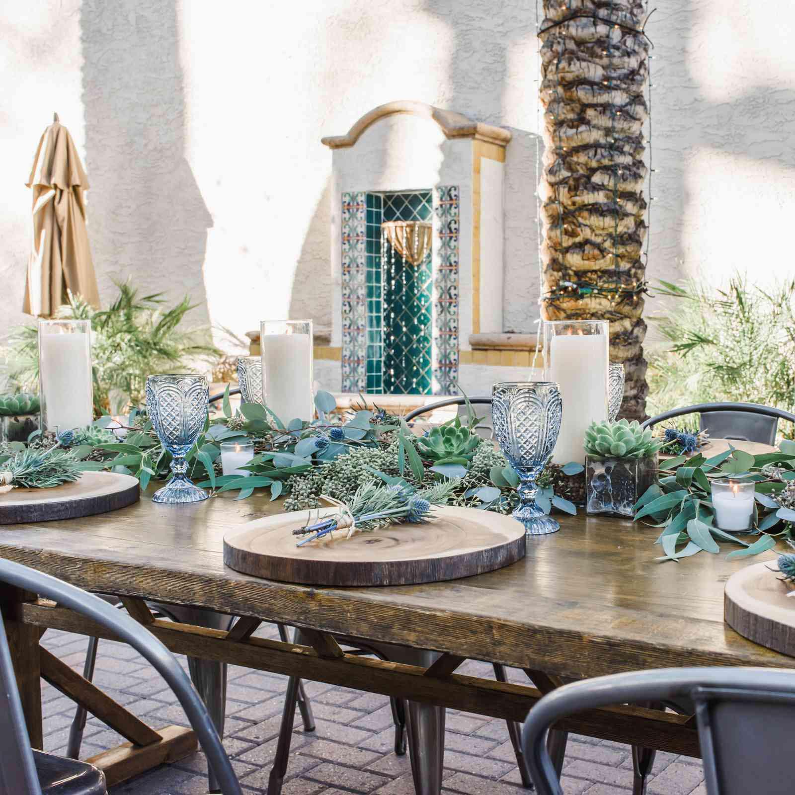 A beautifully set outdoor dining table with wooden boards, candles, greenery, and blue glasses.