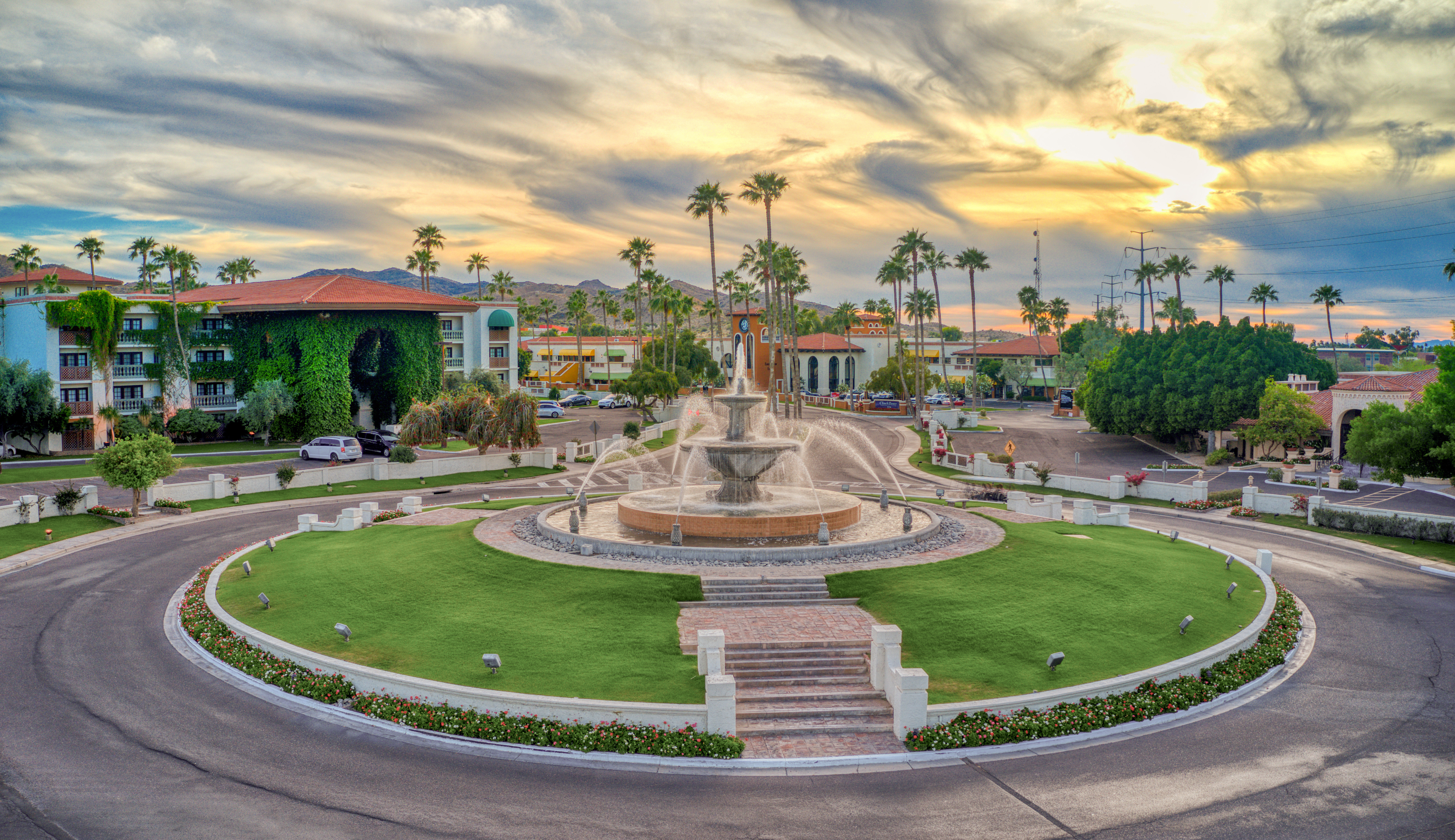 A large fountain in a circular courtyard with a grassy area, palm trees, and a parking lot in the background at sunset.