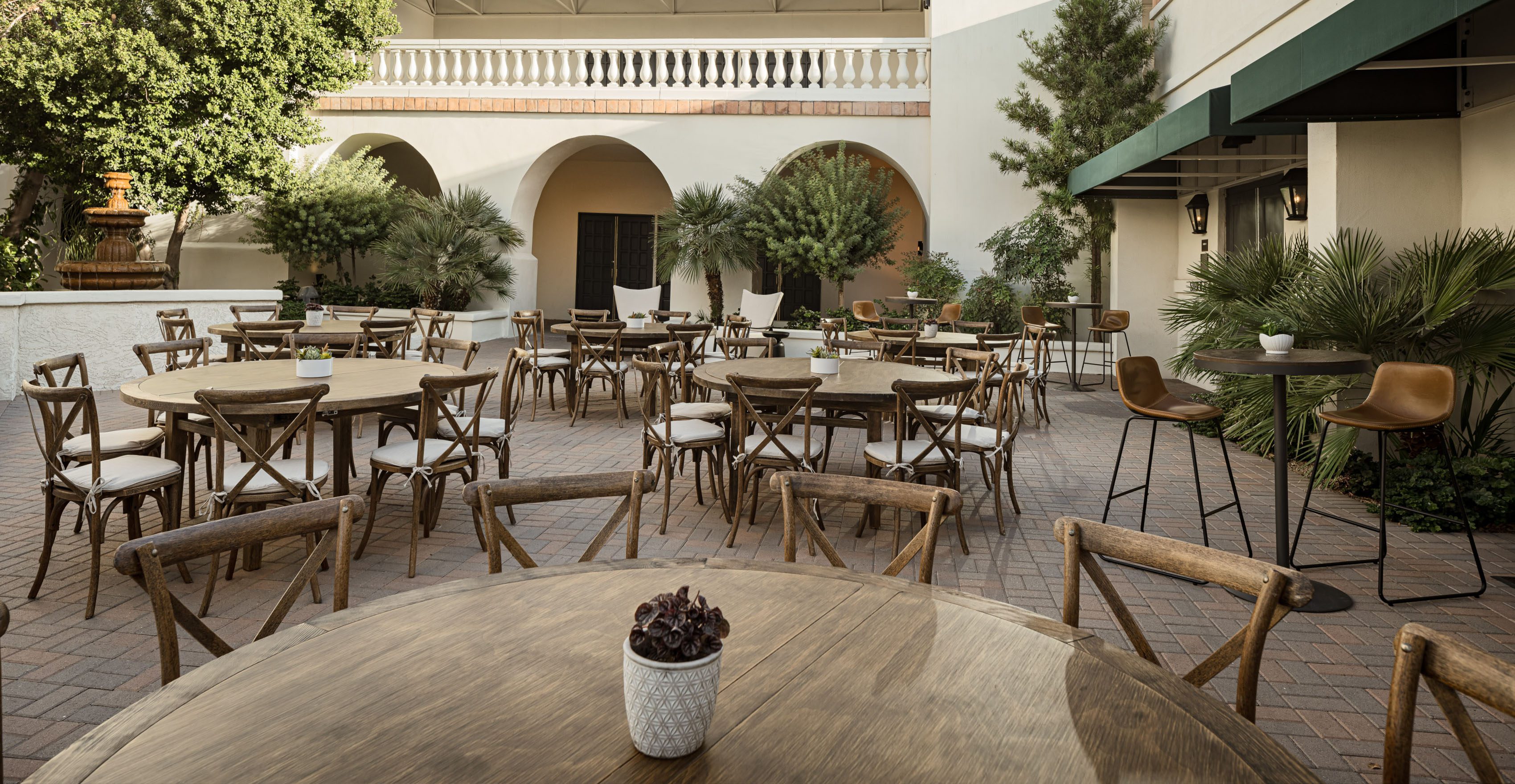 An outdoor dining area with wooden tables and chairs, brick flooring, and a building with arches and a balcony in the background.