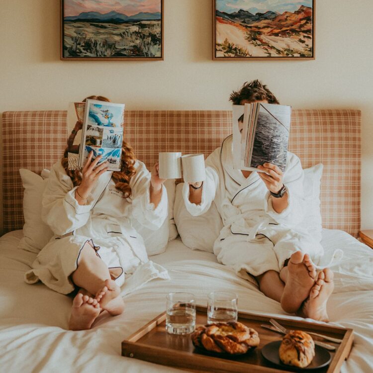 A couple in white robes sitting on a bed with a breakfast tray, reading newspapers.