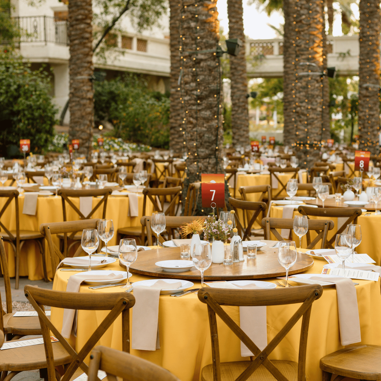 A large outdoor dining area with yellow tables and wooden chairs set up for an event, surrounded by palm trees and string lights.